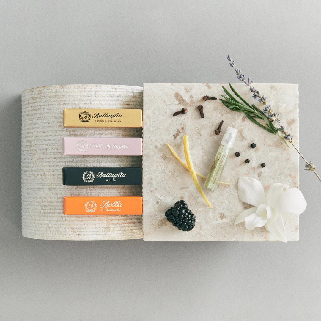 A banner with Battaglia sample boxes on top of a marble stand with the following ingredients next to it: lemon peel, blackberry, white orchid, rosemary, lavender, cloves, and pepper.