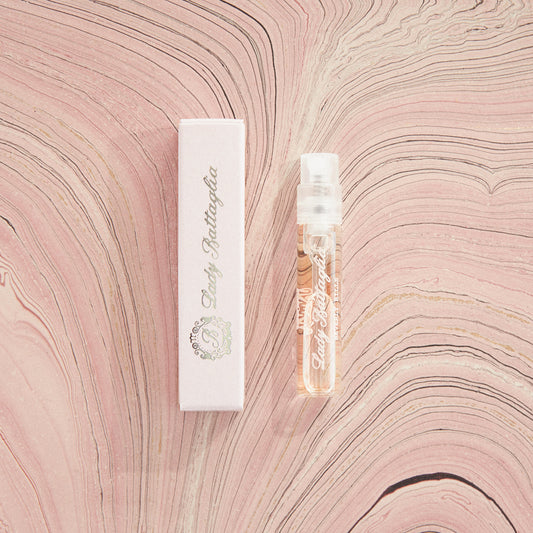 A photo of the Lady Battaglia fragrance sample by Battaglia. The sample is standing upright next to the pink box it is packaged in against a pink marbled paper background.