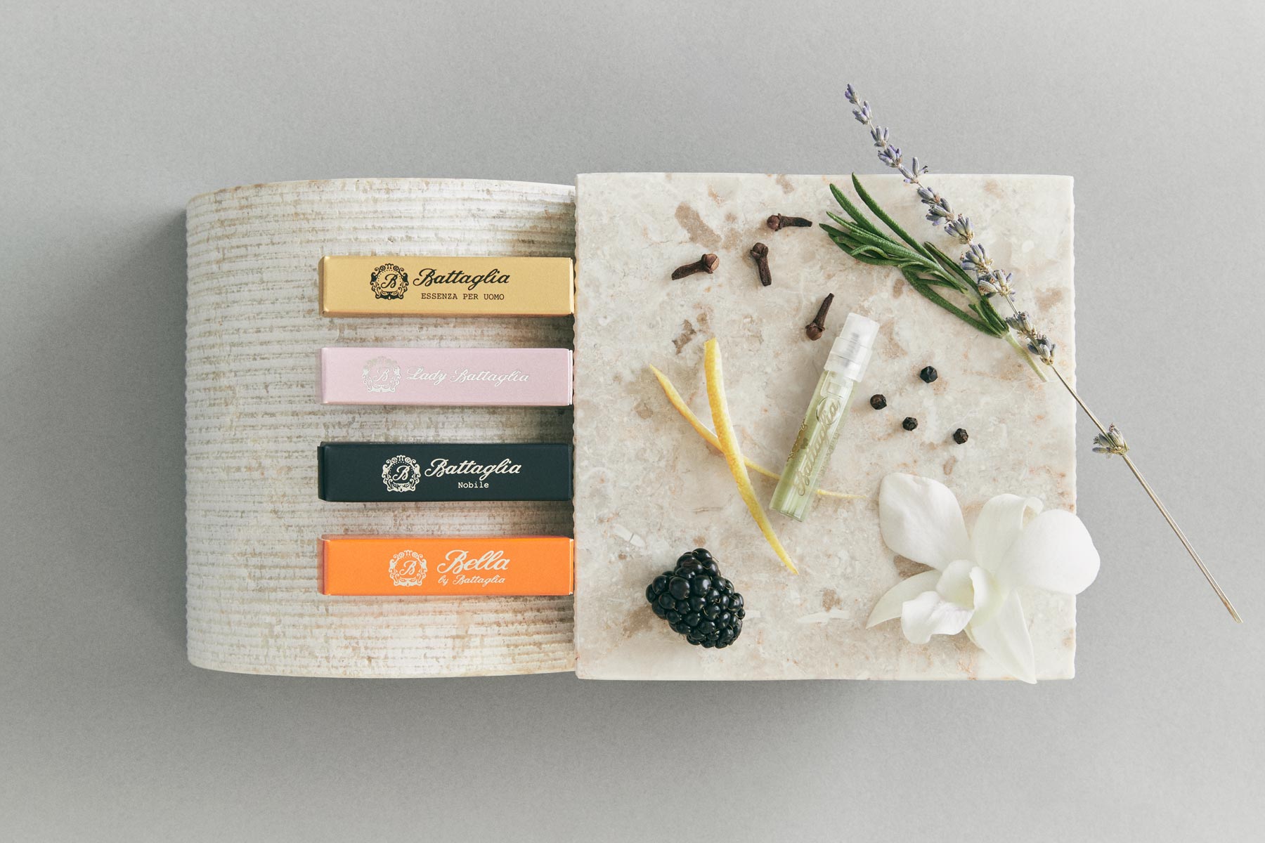 A banner with Battaglia sample boxes on top of a marble stand with the following ingredients next to it: lemon peel, blackberry, white orchid, rosemary, lavender, cloves, and pepper. 