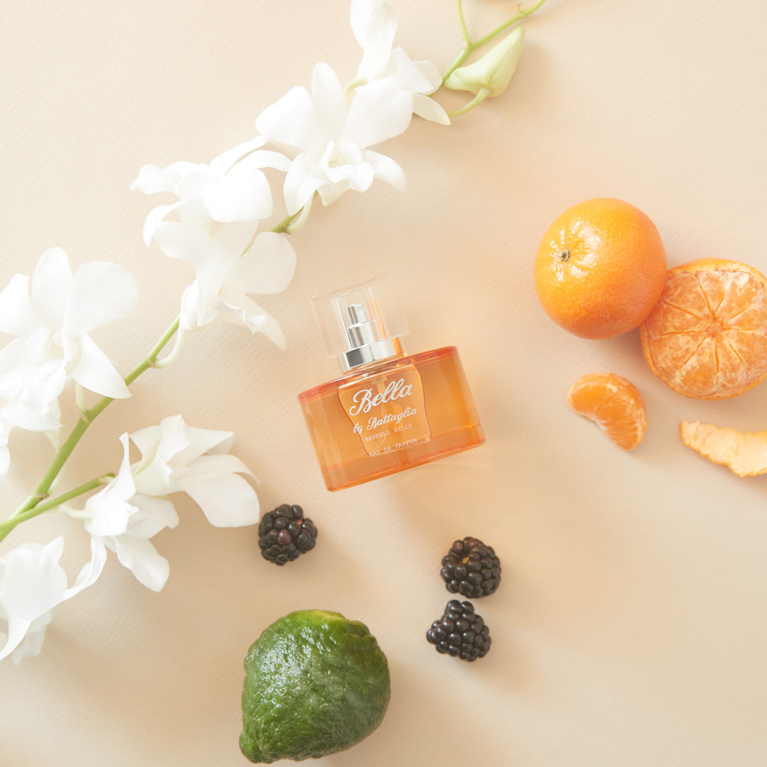 A photo of the Bella fragrance sample by Battaglia. The sample is laying on a flat surface with a light orange background with flowers, citrus, and blackberries spread across the surface.