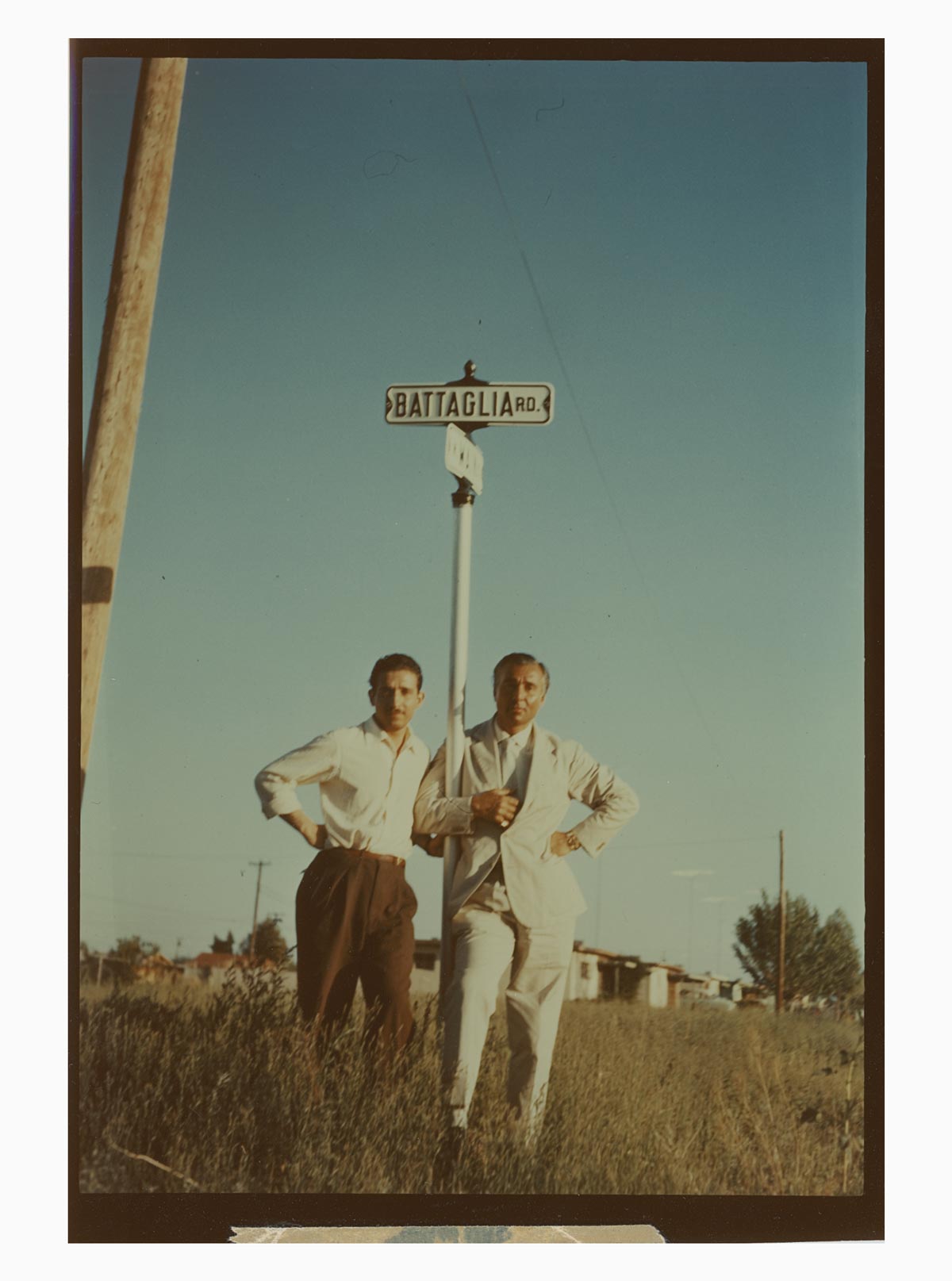 A photo of Battaglia's founder, Giuseppe Battaglia, with a friend standing next to a sign that says Battaglia Rd. Both men look like they are in a grassy field. 