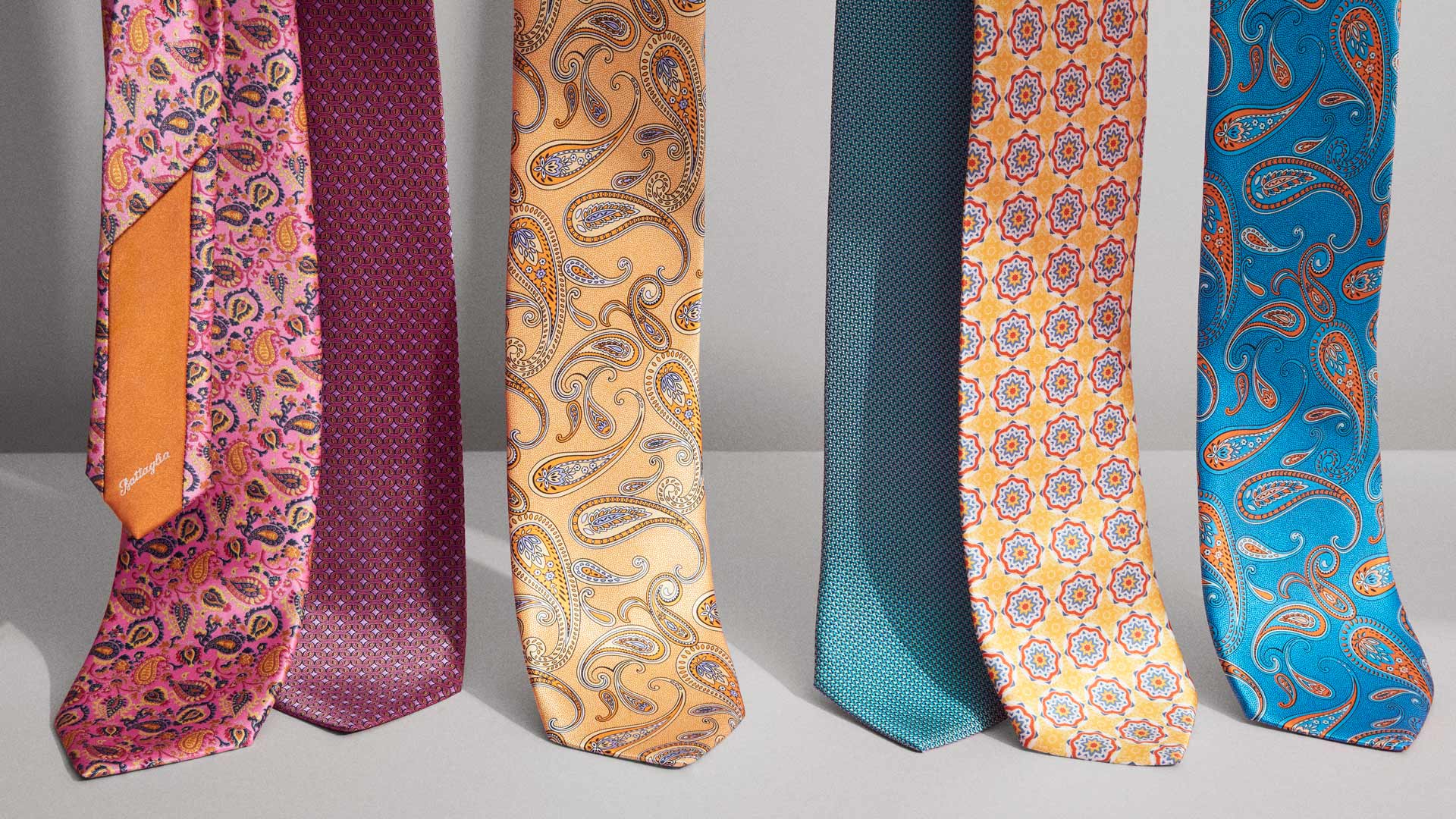 A set of seven colorful Battaglia ties on a grey background