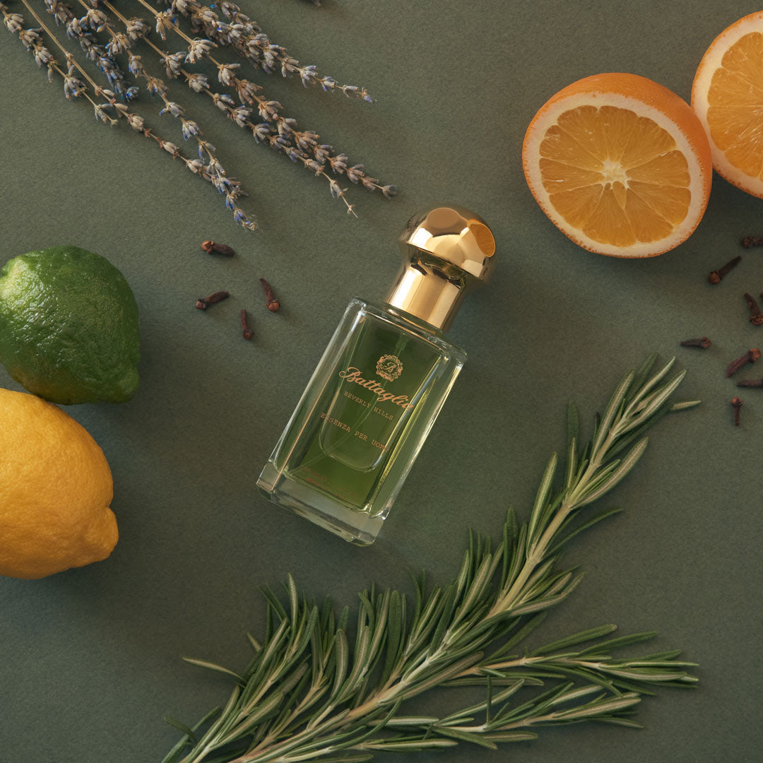 A photo of the Essenza per Uomo fragrance by Battaglia. The sample is laying on a flat surface with a dark green background with citrus, cloves, and rosemary spread across the surface.