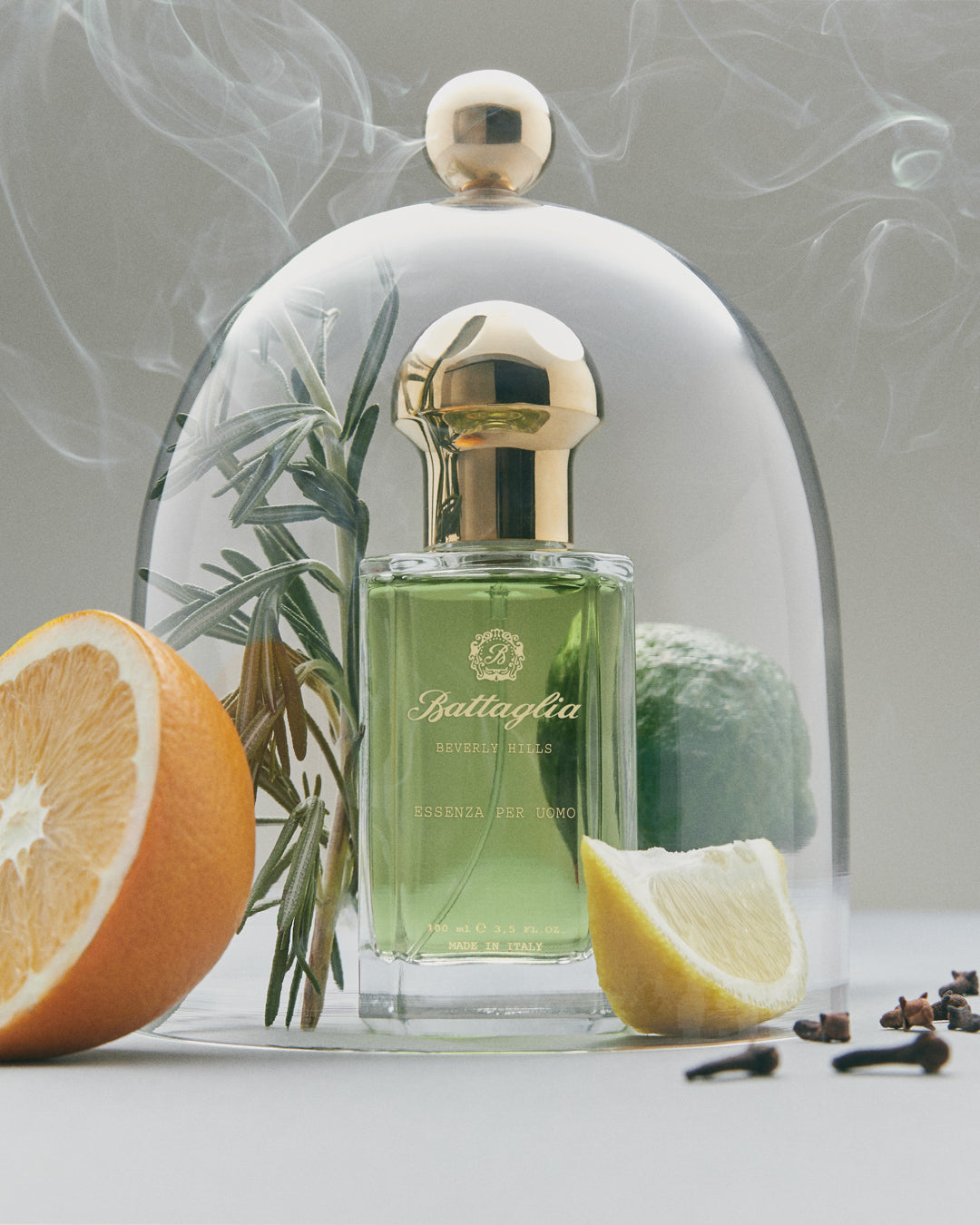 A green bottle of Essenza per Uomo fragrance enclosed in a glass cloche and surrounded by rosemary, bergamot, lemon, cloves, and an orange. 