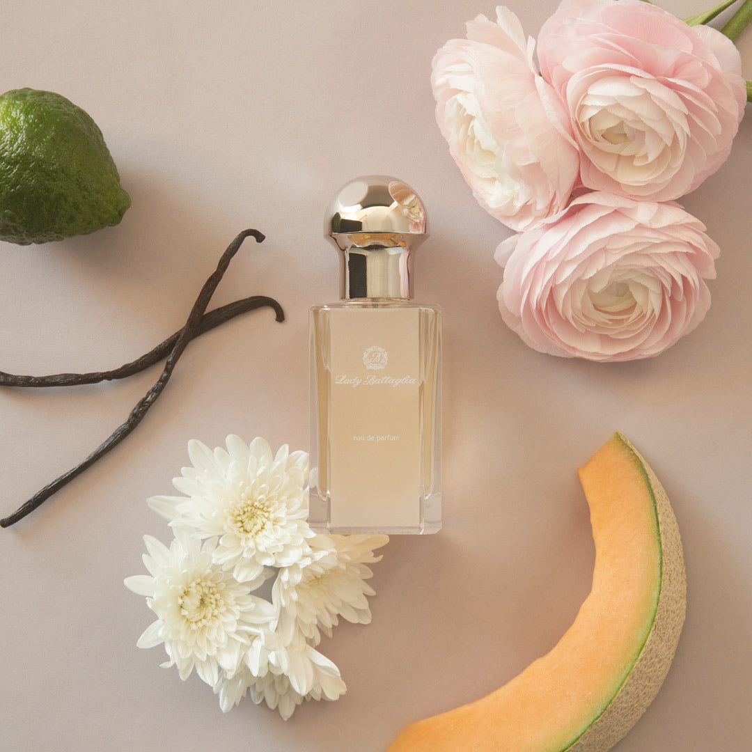 A photo of the Lady Battaglia fragrance by Battaglia. The sample is laying on a flat surface with a light pink background with flowers, citrus, and vanilla bean spread across the surface.