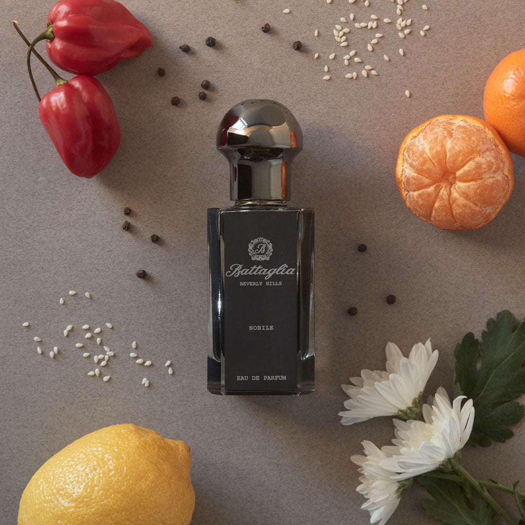 A photo of the Nobile fragrance by Battaglia. The sample is laying on a flat surface with a dark grey background with flowers, citrus, and peppers spread across the surface.