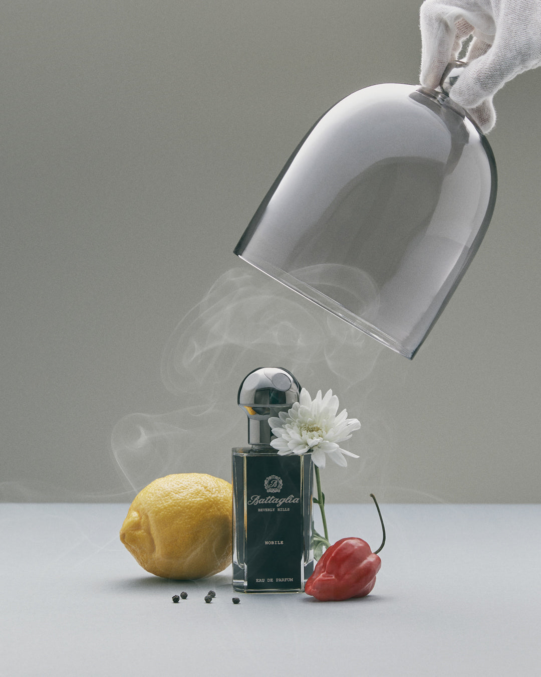A bottle of Nobile fragrance surrounded by a lemon, pepper, white flower, and chili pepper with smoke and a glass cloche.