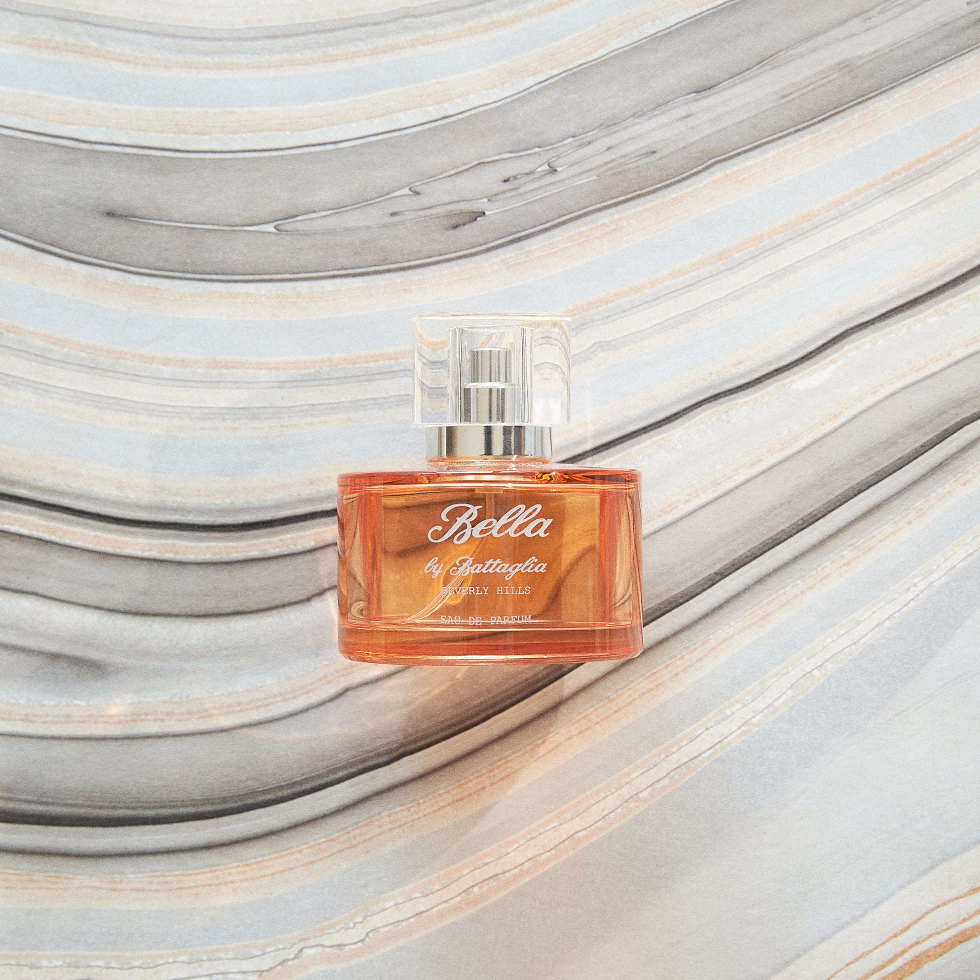 A photo of the Bella fragrance by Battaglia against a grey marbled paper background. The fragrance bottle is round and orange with a clear square top.  