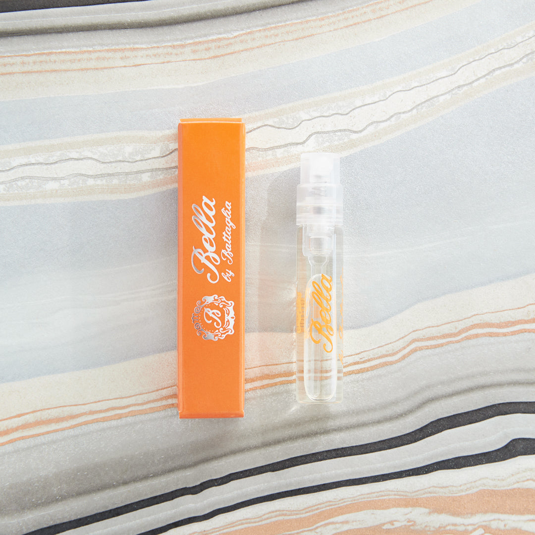 A photo of the Bella fragrance sample by Battaglia. The sample is standing upright next to the orange box it is packaged in against a marbled paper background. 