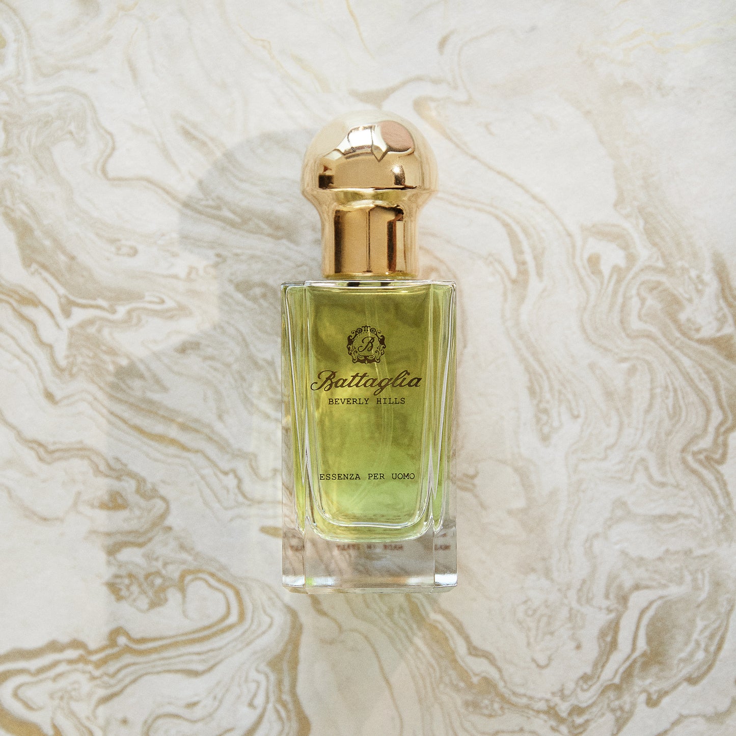 A photo of the Essenza per Uomo fragrance by Battaglia against a gold marbled paper background. The bottle is rectangular with a light green liquid and a gold circular cap.