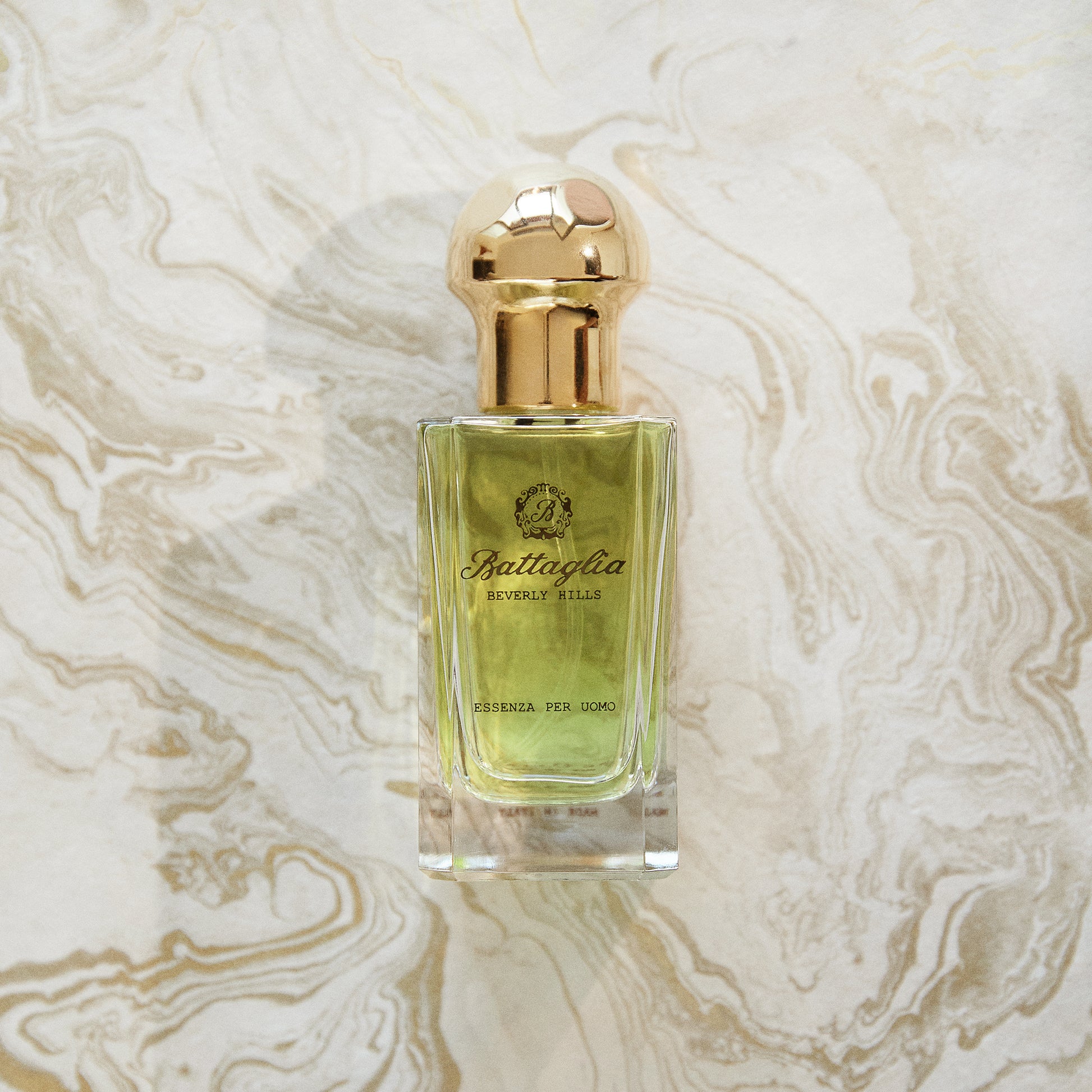 A photo of the Essenza per Uomo fragrance by Battaglia against a gold marbled paper background. The bottle is rectangular with a light green liquid and a gold circular cap.