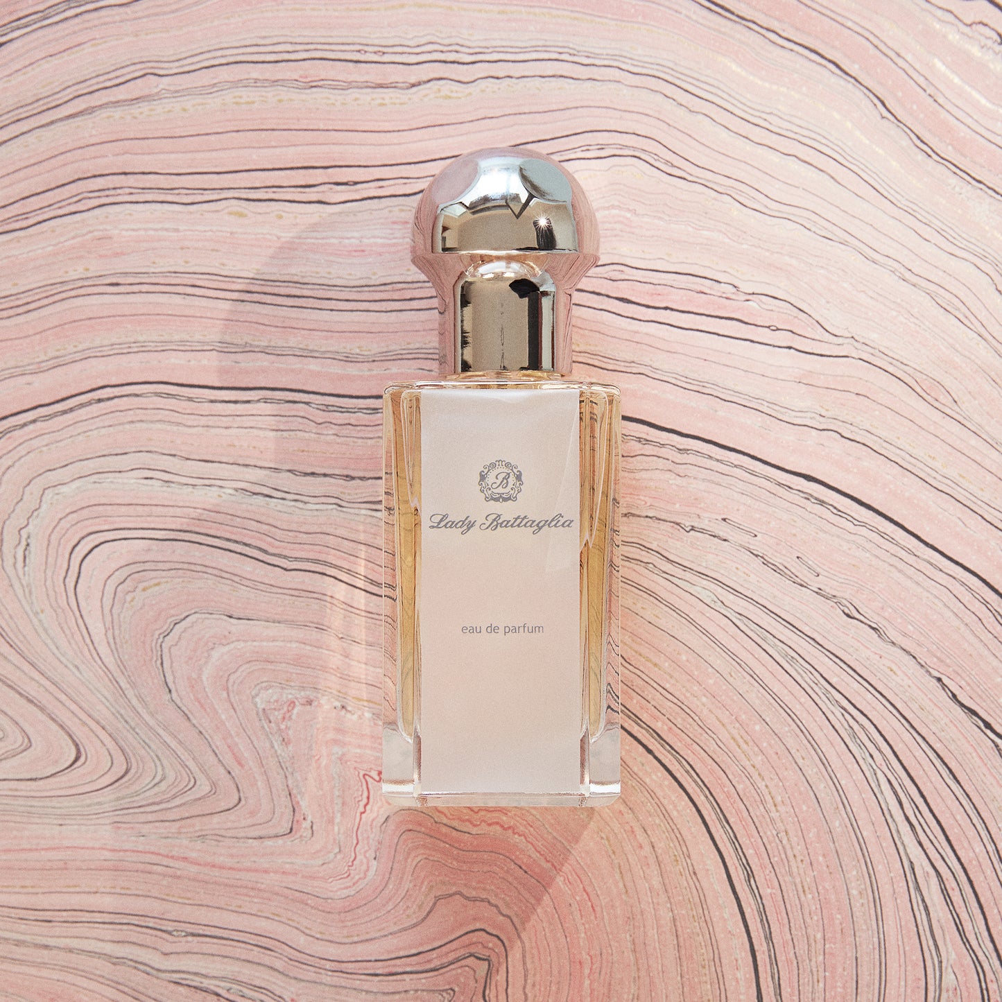 A photo of the Lady Battaglia fragrance by Battaglia against a pink marbled paper background. The bottle is clear and rectangular with a silver circular top. 