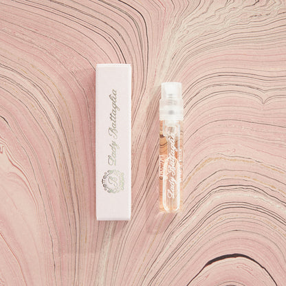 A photo of the Lady Battaglia fragrance sample by Battaglia. The sample is standing upright next to the pink box it is packaged in against a pink marbled paper background. 