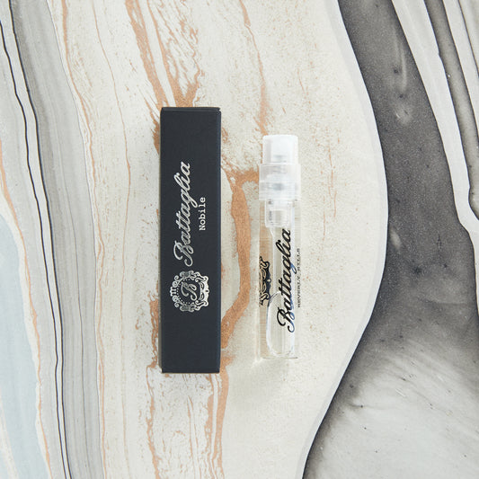 A photo of the Nobile fragrance sample by Battaglia. The sample is standing upright next to the black box it is packaged in against a marbled paper background.